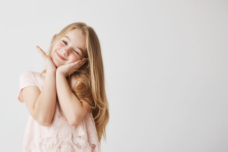 beautiful little blonde girl smiles winking posing touching face with her hands pink cute dress child looking happy delighted copy space